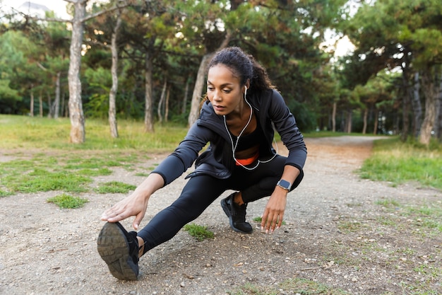 Portrait of fitness african american woman 20s wearing black tracksuit doing exercises, and stretching her legs in green park