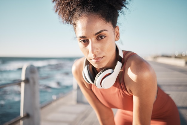 Photo portrait fitness and black woman tired ocean and runner with headphones focus and determined for goal african american lady female exhausted and athlete with podcast rest or training for health