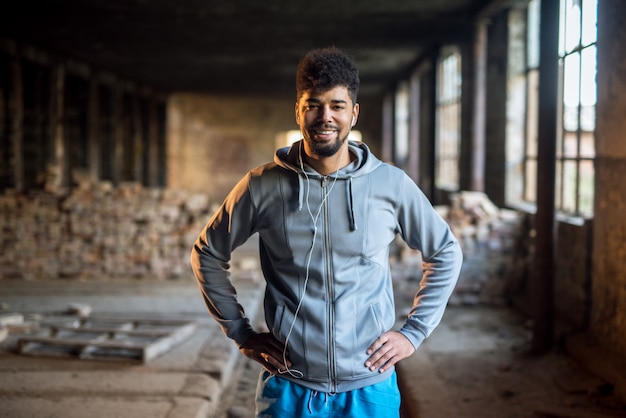 Portrait of focused motivated afro-american young handsome sportive man with earphones standing inside of the abandoned place. Successful young active man portrait.