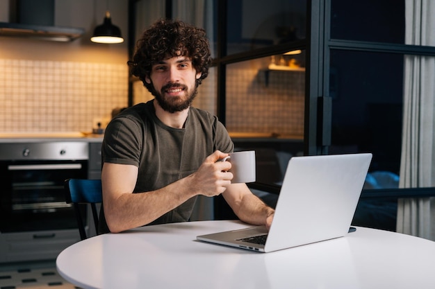 Portrait of friendly bearded young freelancer male holding in hand cup with morning coffee sitting at table with laptop computer looking at camera