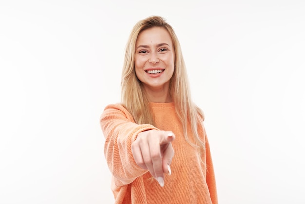 Portrait of friendly blonde girl chooses you points finger at the camera isolated on white background