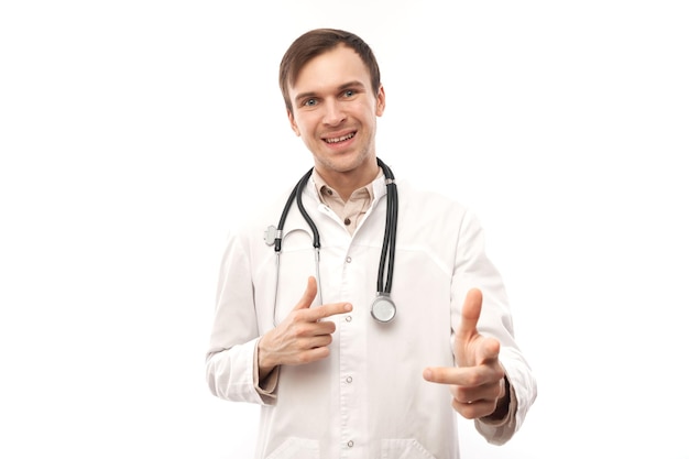 Portrait of friendly positive young male doctor in white medical coat chooses you points finger at camera isolated on white backgroundxA