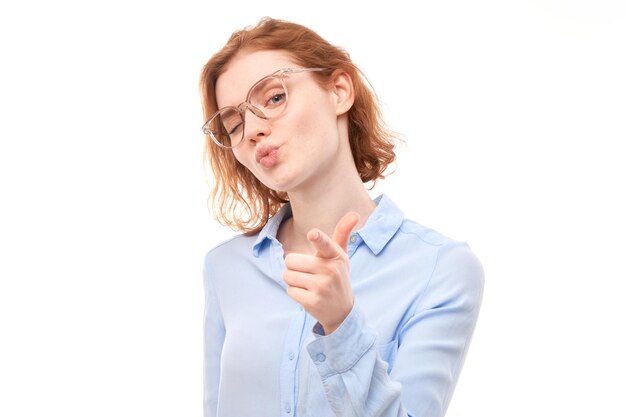 Portrait of friendly redhead girl chooses you points finger at the camera isolated on white background