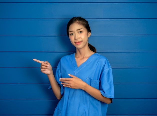 Portrait of friendly smiling confident Asian woman doctor or nurse in blue scrubs suit pointing fingers left showing on blue wooden wall background for advertising about healthcare and medical