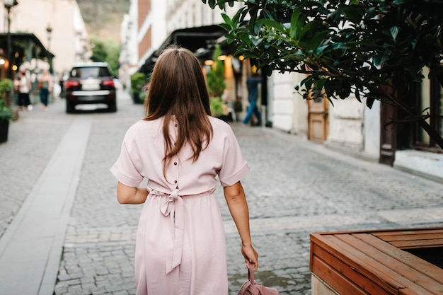 Portrait from back of elegant girl with long hair walking on street near old building on background town Young beautiful girl in stylish and with a fashionable bag