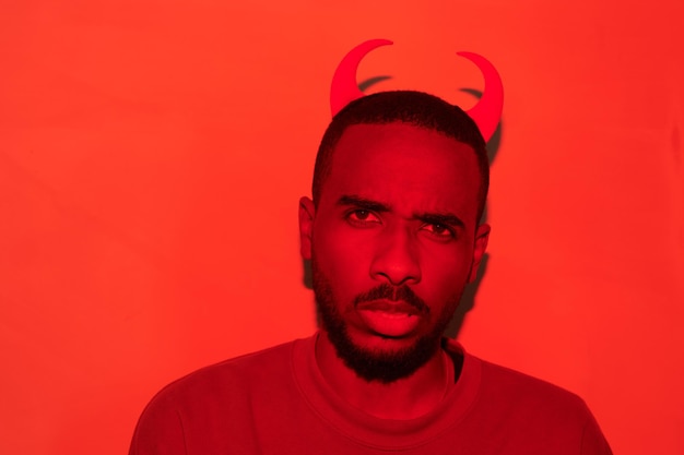 Portrait of frowning puzzled young africanamerican man in devil horns headband standing in red room