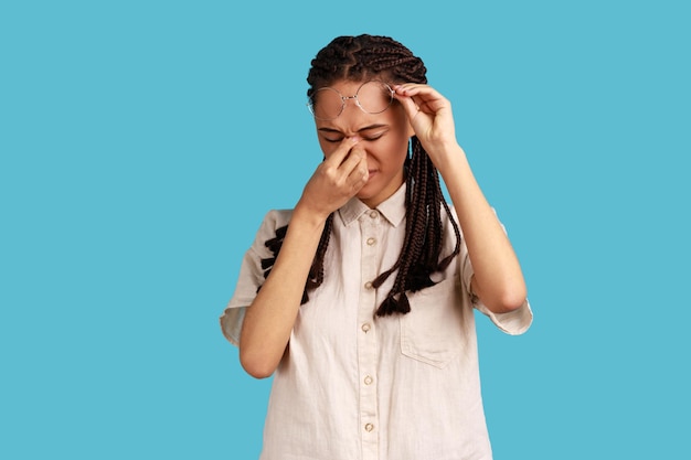 Portrait of frustrated woman with black dreadlocks touching closed eyes, crying from depression, grief or pain, feeling hopeless, wearing white shirt. Indoor studio shot isolated on blue background.