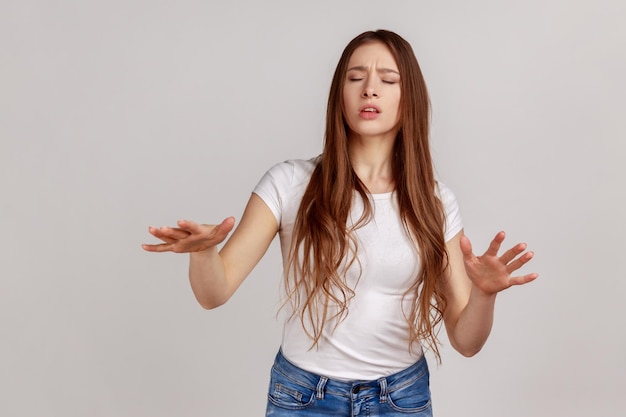 Portrait of frustrated woman with dark hair walking with closed eyes stretching hands to search way in darkness vision problems wearing white Tshirt Indoor studio shot isolated on gray background