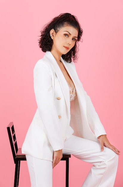 Portrait full body studio shot of Asian sexy curly hairstyle successful businesswoman in white fashionable casual suit with lace lingerie crop top bra sitting on tall chair posing on pink background