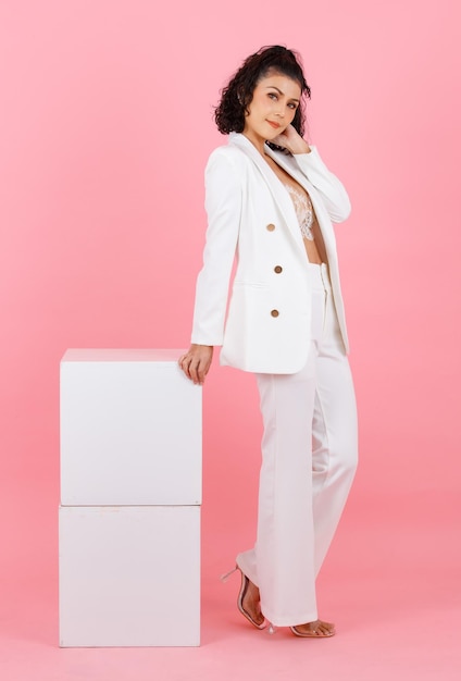 Portrait full body studio shot of Asian sexy curly hairstyle successful businesswoman in white fashionable casual suit with lace lingerie crop top bra standing leaning on two boxes on pink background