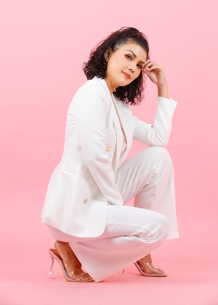 Portrait full body studio shot of Asian young sexy curly hairstyle businesswoman in white fashion casual suit high heels sitting smiling look at camera kneel down posing on floor on pink background