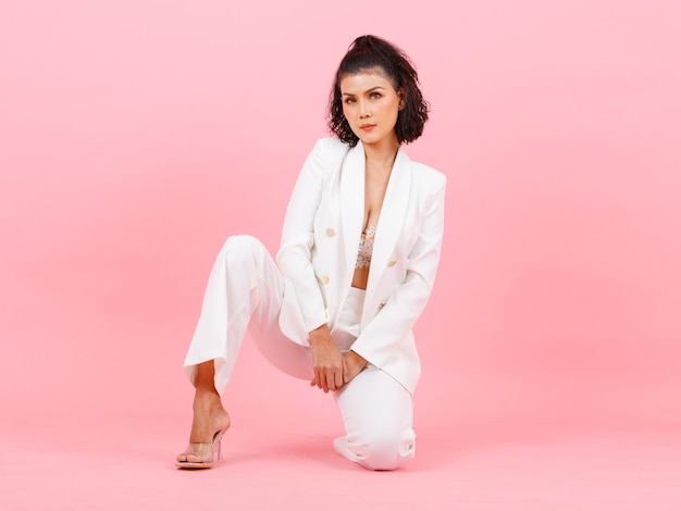 Portrait full body studio shot of Asian young sexy curly hairstyle businesswoman in white fashion casual suit high heels sitting smiling look at camera kneel down posing on floor on pink background