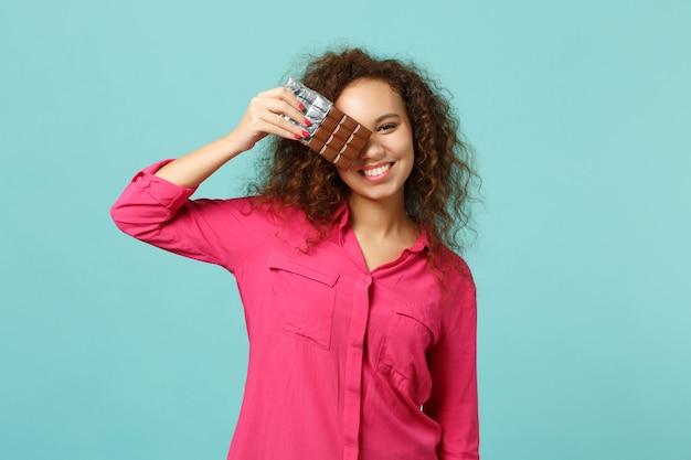 Portrait of funny african girl in casual clothes covering eye with chocolate bar isolated on blue turquoise wall background in studio. People sincere emotions, lifestyle concept. Mock up copy space.