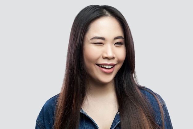 Portrait of funny beautiful brunette asian young woman in casual blue denim jacket with makeup standing with toothy smile winking and looking at camera. studio shot, isolated on light grey background.