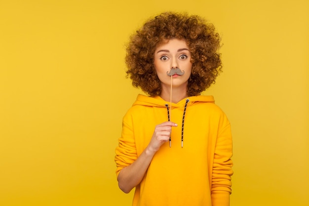 Portrait of funny curlyhaired woman in urban style hoodie keeping fake paper mustache on lips and looking at camera with amazed humorous expression indoor studio shot isolated on yellow background