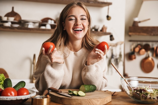 portrait of funny cute woman wearing casual clothes laughing and holding tomatoes in cozy kitchen