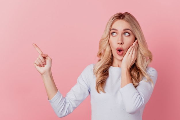 Portrait of funny girlish lady point finger blank space look novelty open mouth palm cheek on pink background