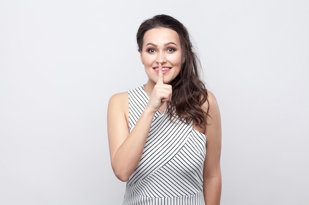 Portrait of funny happy beautiful young brunette woman with makeup and striped dress standing, smiling and looking at camera with secret sign. indoor studio shot, isolated on grey background.