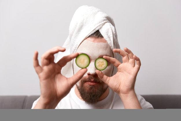 Portrait of funny happy man in white tshirt applying cucumber slices over clay mask on his face. Self care morning spa procedure. Beauty treatment concept