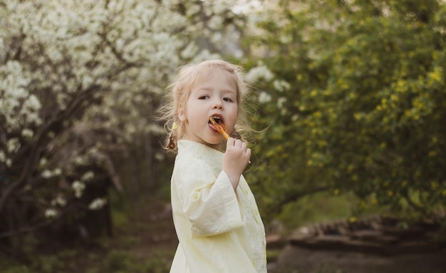 Portrait of funny little girl in yellow dress sucking candy on the background flowering garden