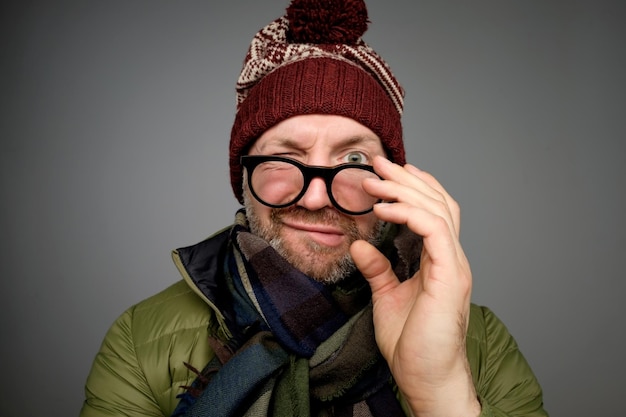 Portrait of a funny mature man in warm winter clothes looking at camera through glasses over gray background