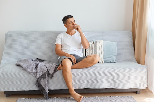 Portrait of funny pensive handsome young adult man wearing white shirt and jeans short, looking away with thoughtful facial expression, putting finger into nose, posing indoor while sitting on sofa.