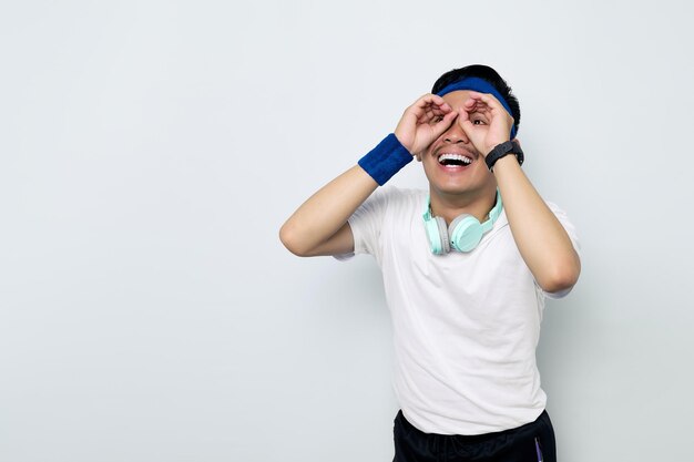 Portrait of funny young Asian sportman in blue headband and sportswear white tshirt with headphones showing eyeglasses binoculars isolated on white background Workout sport concept