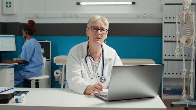 Portrait of general practitioner using laptop at health care checkup visit, working on appointment paperwork. Female physician looking at diagnosis results on medical system online.