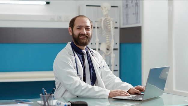 Portrait of general practitioner working on laptop to plan checkup visit, wearing white coat and stethoscope in medical cabinet. Male physician using computer to cure sick patients.