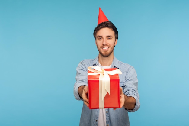 Portrait of generous kind cheerful man with party cone hat giving gift, congratulating on birthday and offering surprise, holiday bonus wrapped in box. indoor studio shot isolated on blue background