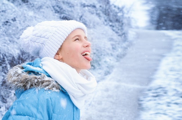 Portrait of a girl in blue winter clothes catching snowflakes with her tongue in the park.