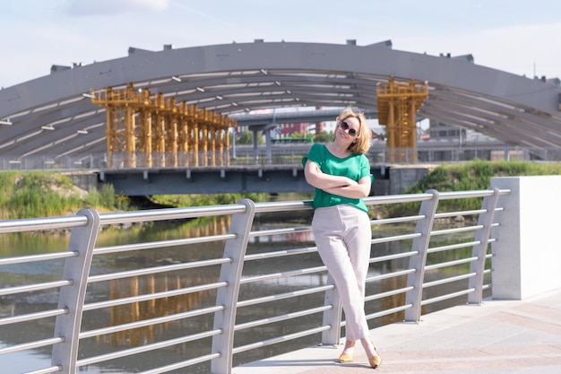 A portrait of a girl in a green blouse trousers and sunglasses against background of unfinished urban architecture