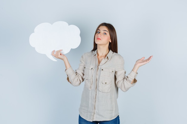 Portrait of girl holding speech bubble, spreading palm aside in shirt and looking indecisive