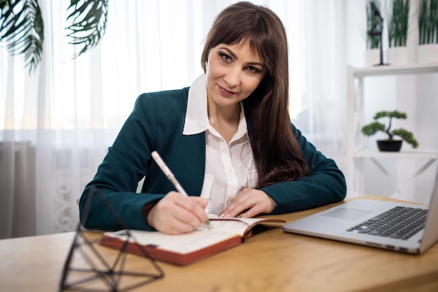 Portrait of a girl in a jacket and shirt sits at a table and writes notes in a notebook from a laptop