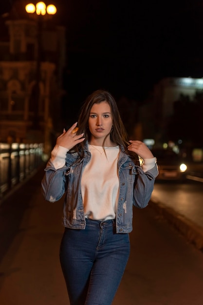 Portrait of girl student walking on night street Stylish attractive young woman in denim jacket evening city