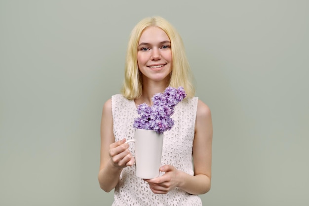 Portrait of girl teenager with flowers on green background. Beautiful female blonde holding lilac in cup. Youth, spring, beauty, cosmetology, health concept