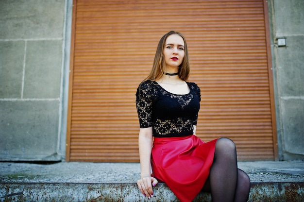 Portrait of girl with black choker necklace on her neck and red leather skirt against orange shutter.