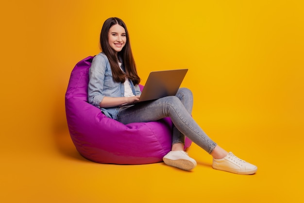 Portrait of girl with laptop sit beanbag look camera on yellow background