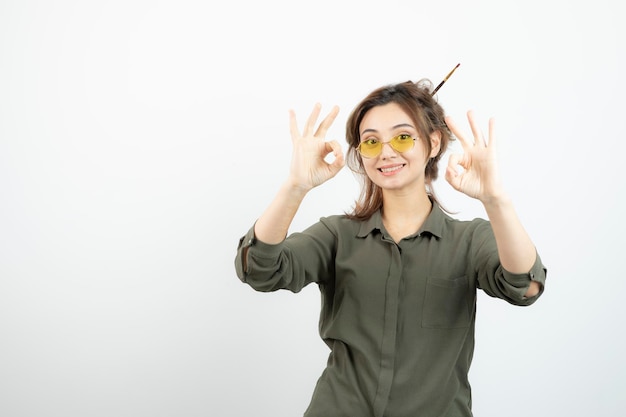 Portrait of girl with messy bun in glasses standing and giving ok sign. High quality photo