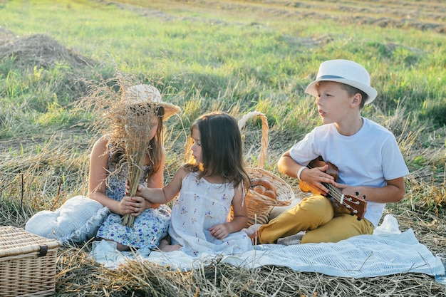 Portrait of glad three kids sitting on blanket in field playing ukulele having picnic with drinks and tasty food