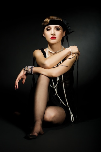 Portrait of a glamorous dark haired model with perfect makeup wearing a luxurious necklace black bodysuit and long top holding her hands behind her head and looking straight ahead