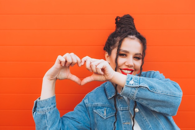 Portrait of a glamorous woman with dreadlocks in a denim suit and with red lips against an orange wall