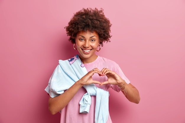 Portrait of good looking Afro American woman shows heart sign and smiles broadly expresses romantic emotions confesses in love cares for someone wears casual t shirt with jumper tied over shoulder