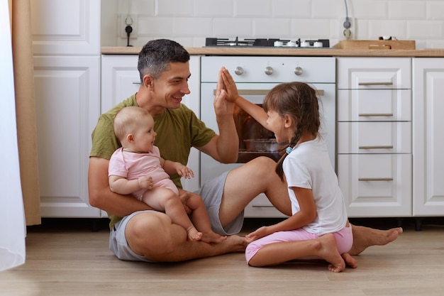 Portrait of good looking father giving five his kid man wearing casual attire sitting on floor with his daughters and baking cookies in oven people spending time together while cooking