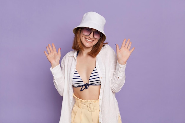 Portrait of good looking pretty young woman wearing panama white shirt and swimming suit posing isolated over purple background standing with raised arms and toothy smile