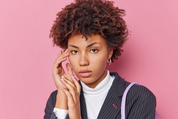 Portrait of good looking woman touches face gently has healthy skin curly hair looks directly at camera with serious calm expression dressed in elegant formal clothes isolated over pink background