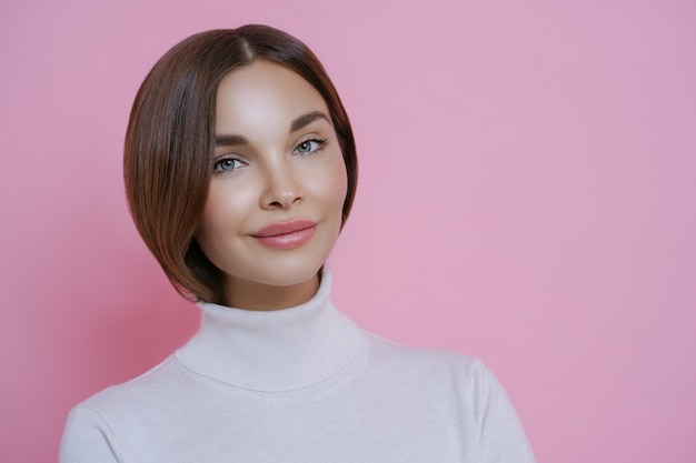 Portrait of good looking young woman with dark hair, minimal makeup, wears casual white turtleneck