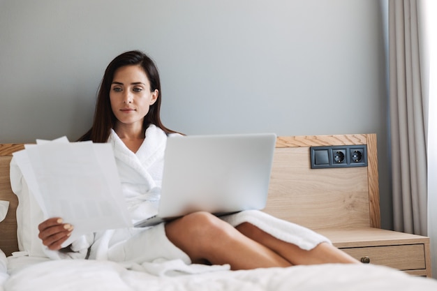 Portrait of gorgeous adult businesswoman in white bathrobe working with paper documents and laptop while lying on bed in apartment