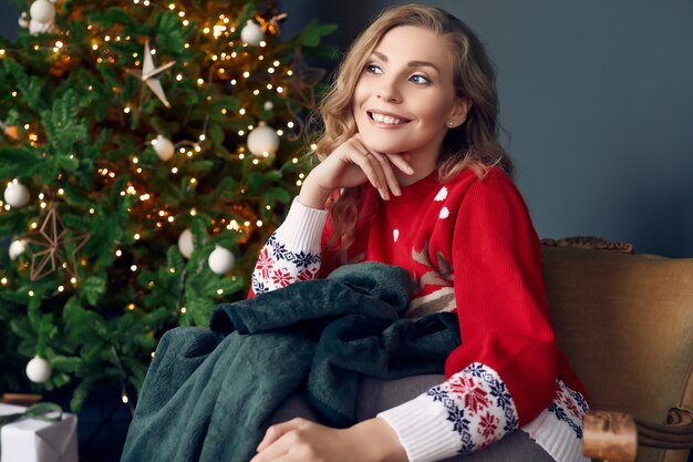 Portrait of gorgeous blonde woman in red sweater chilling under a plaid in the Christmas decorated interior.