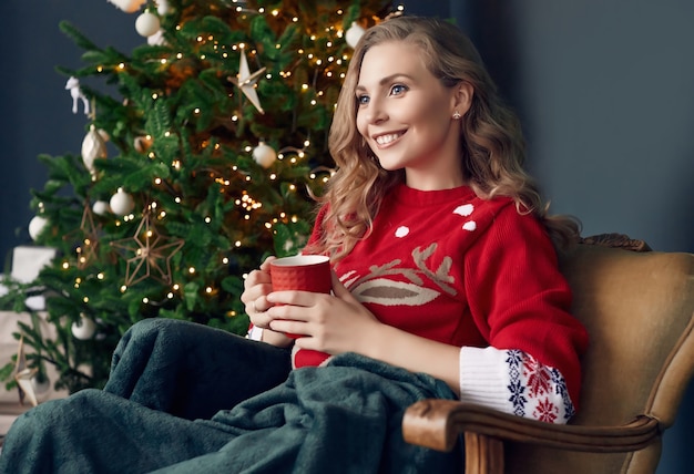 Portrait of gorgeous blonde woman in red sweater chilling under a plaid with cup of coffee in the Christmas decorated interior.